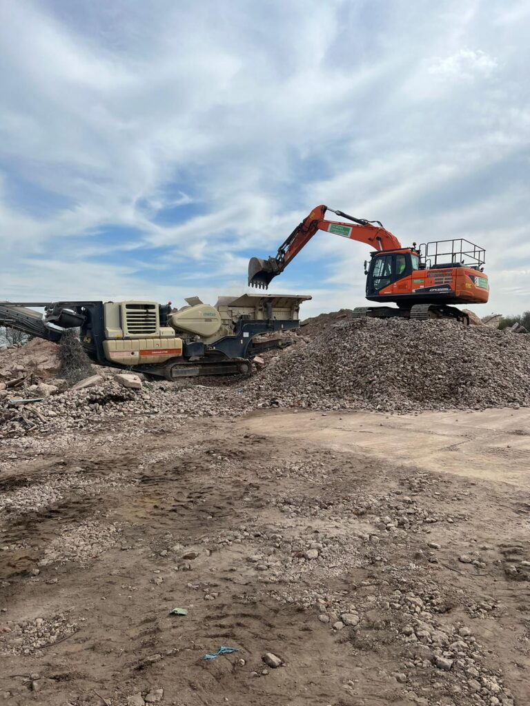 A construction machine crushes concrete while working on a large pile of dirt at a construction site.